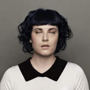 Portrait of a Woman with Blunt Black Bangs and Dark Hair, Eyes Half-closed, Wearing a White Sweater with a Black Collar, Looking Tired or Sleepy.
