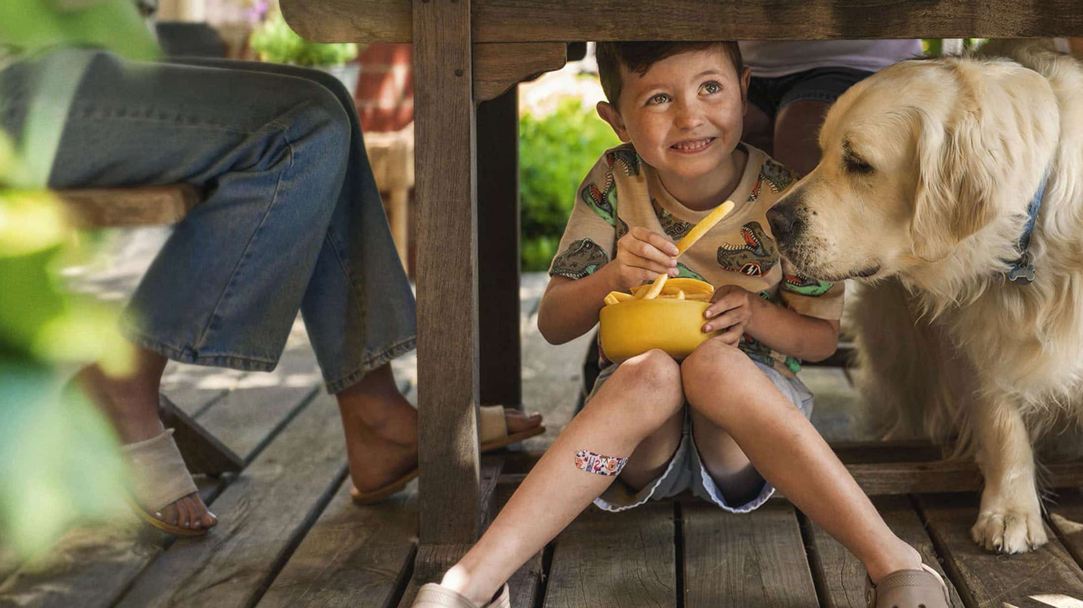 A smiling child under a wooden table sharing fries from a yellow bowl with a golden retriever beside him outdoors.