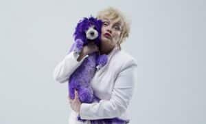 Person in a White Blazer Holds a Purple Fluffy Stuffed Dog Close and Pouts Playfully at the Camera.