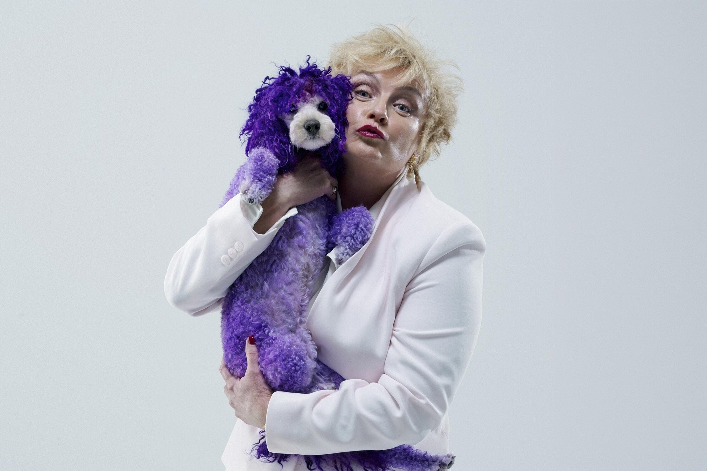 Person in a white blazer holds a purple fluffy stuffed dog close and pouts playfully at the camera.
