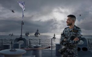 Navy Sailor in Camouflage Uniform Stands on a Ship Deck with Arms Crossed, Looking Toward the Horizon As Helicopters Fly Overhead and Battleships Anchor in a Cloudy Sea.