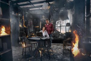 Two Adults Standing in a Charred, Fire-destroyed Living Room Beside a Table with a Laptop, Surrounded by Flames and Smoke.