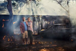 Couple Stands Arm-in-arm in Front of a Burning House with a Smoldering Car Nearby.