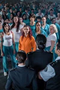 Large Crowd of Inmates in Teal Uniforms Facing Two Guards in the Foreground; a Central Inmate Wears an Orange Jumpsuit Looking Forward.