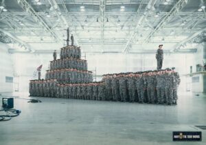 Large Formation of Soldiers in Camouflage Uniforms Inside a Spacious Hangar, with a Pyramid Stack and a Lone Officer Standing to the Right.