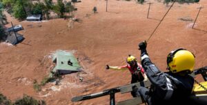 Aerial View of a Flood Rescue: a Helicopter Hoists a Person Across a Red Muddy River While Others Stand on Rooftops in Submerged Houses.