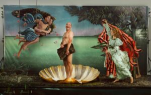 the Staged Scene Shows a Man Standing in a Giant Shell with a Sea Backdrop, Flanked by a Winged Figure and a Woman in a Red Cape and Pale Dress.