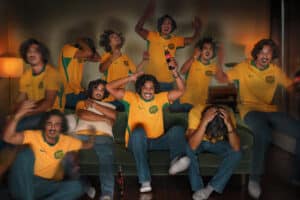 Group of Men in Yellow Brazil Jerseys Cheer and Celebrate on a Sofa, a Coca-cola Bottle Visible on the Table Nearby.