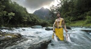 Man in Yellow Waders Standing in a Rocky River, Holding Two Large Fish, with a Forested Riverbank and Mountain in the Background.
