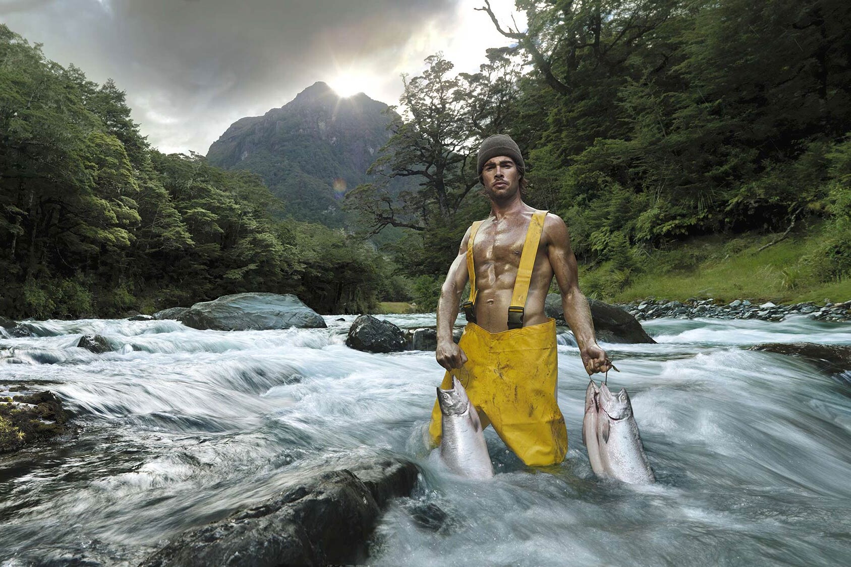 Man wearing yellow waterproof waders stands in a rushing river, holding two fish in each hand.