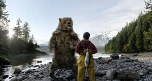 a Hunter in a Plaid Jacket and Yellow Overalls Stands Beside a Giant Standing Bear on a Rocky Lakeshore with Snow-capped Mountains in the Background, Near a Pine Forest.