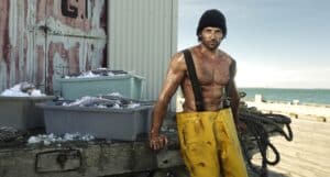 Fisherman in Yellow Waterproof Pants and a Black Knit Cap Stands on a Dock Beside Ice-filled Fish Bins, with the Sea in the Background.