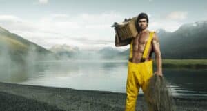 Fisherman in Yellow Waders Carries a Wicker Basket and Fishing Net by a Misty Alpine Lake with Mountains in the Background.