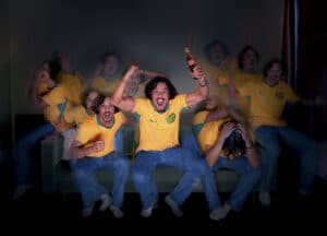 Group of Friends in Matching Yellow Brazil Jerseys Celebrate on a Couch, with One Person Raising a Coca‑cola Bottle Amid Motion Blur.