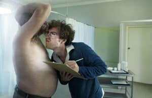 Medical Exam Scene: a Clinician in Glasses Leans in to Inspect a Shirtless Patient’s Chest While Holding a Clipboard in a Clinic Room