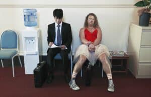 Two Men Waiting: One in a Suit Reading Papers, the Other in a Red Dress with a Wig and Bracelets, Legs Crossed in a Waiting Room.