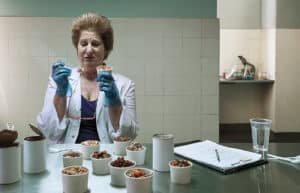a Researcher in a White Lab Coat and Blue Gloves Tastes Samples from Small Cups on a Stainless Table, Clipboard and Glasses Nearby.