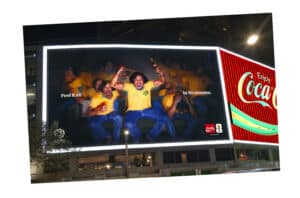 Nighttime Coca-cola Billboard Showing Cheering Men in Yellow Shirts, with the Text 'feel It All' and 'in 90 Minutes' and the Coca-cola Logo.