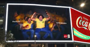 Bright Billboard Featuring Excited Football Fans in Yellow Jerseys, Raising Fists; the Ad Reads 'feel It All in 90 Minutes' with Coca‑cola Branding Nearby.